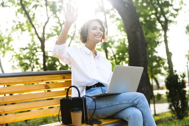 Gülümseyen genç kadın açık havada Street'teki bankta oturmuş dizüstü bilgisayar, el sallayarak kullanarak
