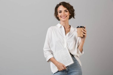 Mutlu bir genç kadının poz fotoğraf izole içme kahve holding gazete gri arka plan üzerinde.