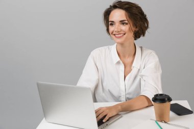 Dizüstü bilgisayar kullanarak masada oturan gri duvar arka plan üzerinde izole görüntüsünü poz genç mutlu neşeli iş kadını.