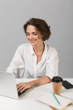 Dizüstü bilgisayar kullanarak masada oturan gri duvar arka plan üzerinde izole görüntüsünü poz genç mutlu neşeli iş kadını.