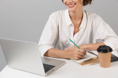 Dizüstü bilgisayar kullanarak masada oturan gri duvar arka plan üzerinde izole genç neşeli iş kadın poz kırpılmış fotoğrafı.