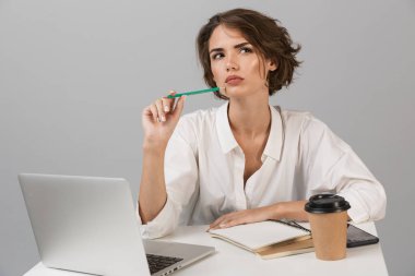 Görüntü dizüstü kullanarak masada oturan gri duvar arka plan üzerinde izole iş ciddi düşünceli genç kadının poz.