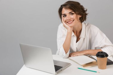 Dizüstü bilgisayar kullanarak masada oturan gri duvar arka plan üzerinde izole görüntüsünü poz genç mutlu neşeli iş kadını.