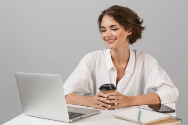 Dizüstü bilgisayar kullanarak masada oturan gri duvar arka plan üzerinde izole görüntüsünü poz genç mutlu neşeli iş kadını.