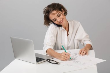 Telefonla konuşurken laptop çizim kullanarak masada oturan gri duvar arka plan üzerinde izole görüntüsünü poz genç iş kadını.