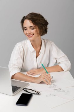 Dizüstü çizim kullanarak masada oturan gri duvar arka plan üzerinde izole görüntüsünü poz genç iş kadını.