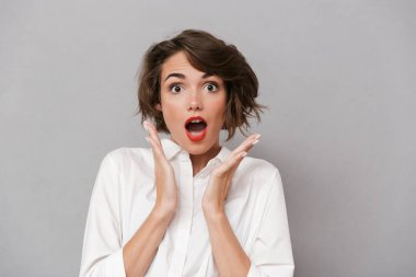 Şok bir genç kadının portresi kameraya bakarak gri arka plan üzerinde izole ayakta beyaz gömlek giymiş