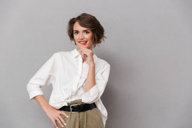 Dalgın bir genç kadının portresi kameraya bakarak gri arka plan üzerinde izole ayakta beyaz gömlek giymiş