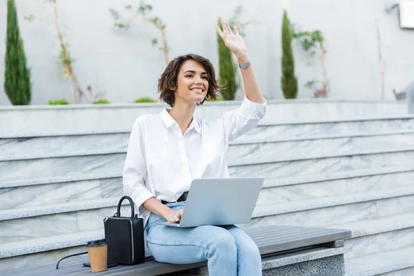 Gülümseyen genç kadın açık havada Street'teki bankta oturmuş elini sallayarak, dizüstü bilgisayar kullanarak