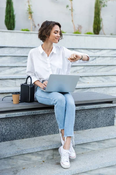 Gülümseyen genç kadın açık havada Street'teki bankta oturmuş dizüstü bilgisayar, zaman kontrol kullanarak