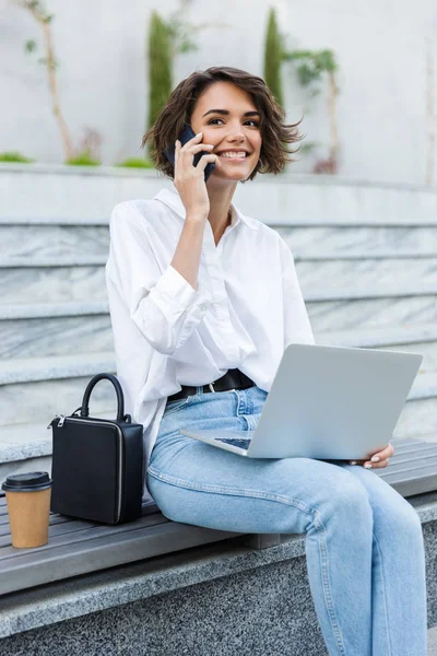 Gülümseyen genç kadın açık havada Street'teki bankta oturmuş dizüstü bilgisayar, cep telefonu üzerinde konuşurken kullanarak