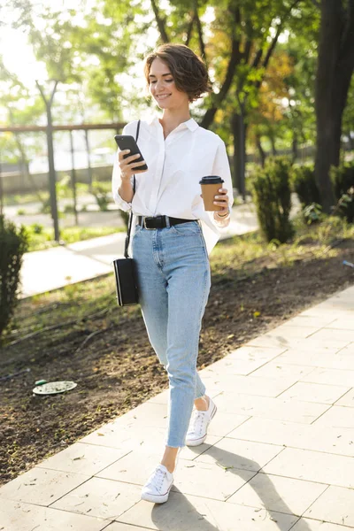 Mujer Joven Sonriente Caminando Por Calle Sosteniendo Teléfono Móvil Bebiendo —  Fotos de Stock