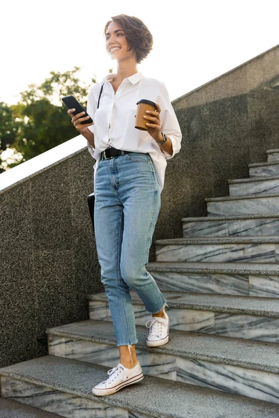 Mujer Joven Sonriente Caminando Abajo Calle Usando Teléfono Móvil Bebiendo —  Fotos de Stock