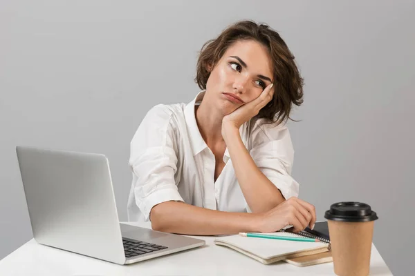 照片中 一个无聊的疲惫的年轻女子坐在桌子旁 在笔记本电脑附近的灰色背景中隔离 — 图库照片