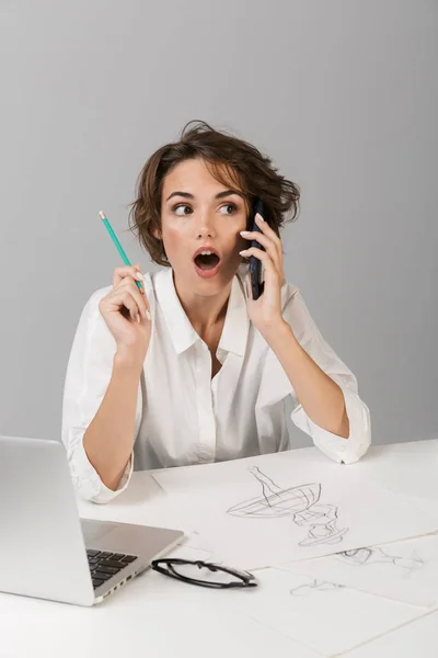 年轻的商业形象震惊的妇女摆在灰色的墙壁背景孤立的妇女坐在桌子上 用笔记本电脑画画电话 — 图库照片