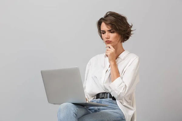 照片中 一个有思想的年轻女子坐在凳子上 站在灰色背景上 拿着笔记本电脑 — 图库照片
