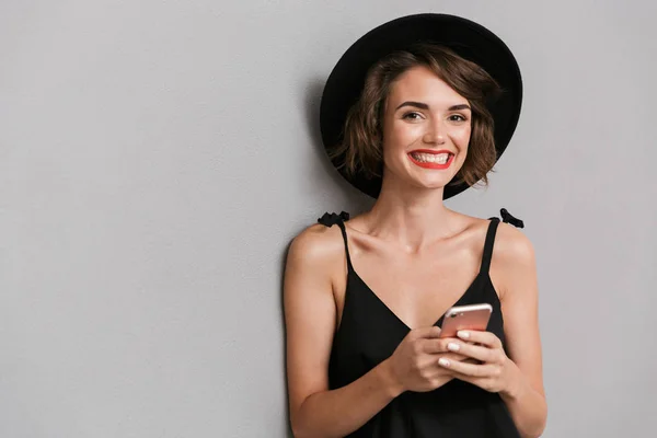 照片中 20岁的快乐女子穿着黑色礼服 头戴帽子 对着镜头微笑 同时拿着在灰色背景上孤立的智能手机 — 图库照片