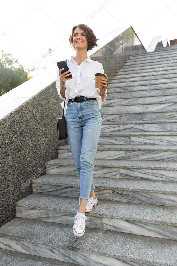 Cheerful young woman walking downstairs at the street, using mobile phone, drinking coffee