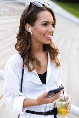 Mutlu genç kadın parkta vakit, yürüme, limonata içmeye, cep telefonunuzun ve