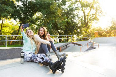 Mutlu iki kız arkadaş Sisters park açık havada içinde oturan silindirler üzerinde görüntü almak bir selfie cep telefonuyla.