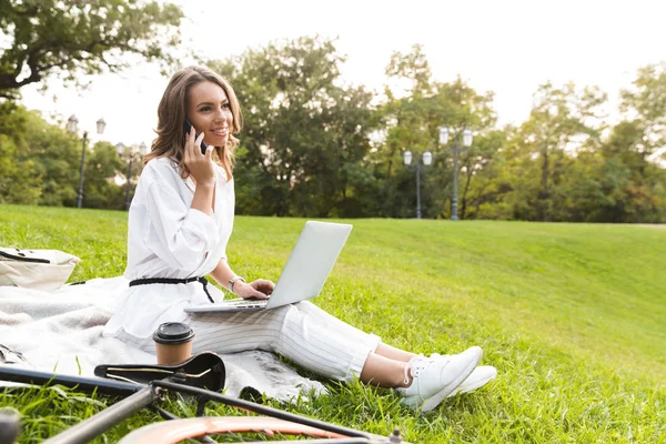 かなり若い女性自転車公園で時間を費やして 草の上に座って ラップトップ コンピューターを使用して 携帯電話で話しています — ストック写真