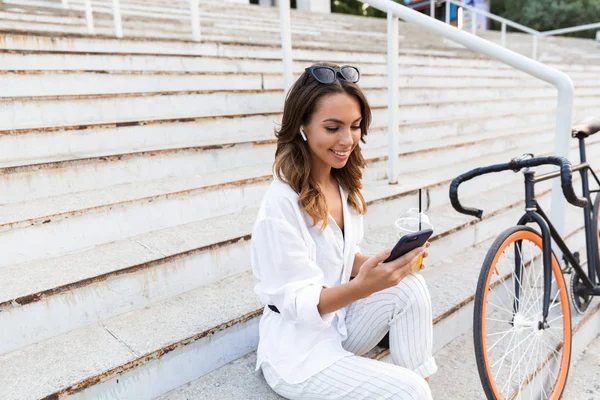 Happy young woman spending time at the park, sitting with bicycle on stairs, drinking lemonade, using mobile phone