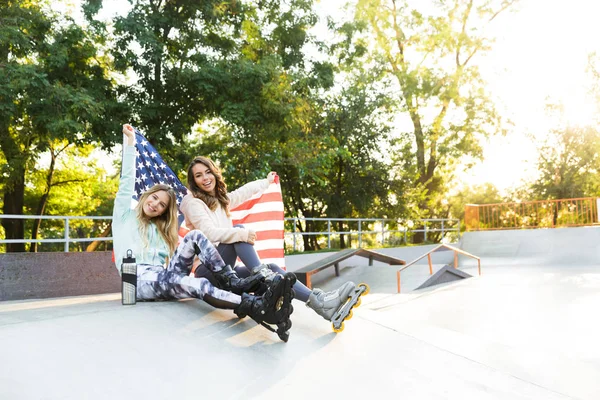 愉快的二个女孩朋友姐妹的图片在滚轮坐在公园户外拿着旗子 Usa — 图库照片
