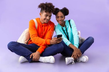 Afro-Amerikan erkek ve kadın bacakları ile katta otururken akıllı telefon ve kulaklık kullanarak sırt çantaları giyen izole mor arka plan üzerinde geçti