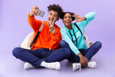 Afrika kökenli Amerikalı adam ve kız bacakları ile katta oturan izole mor arka plan üzerinde çarpı işareti iken selfie fotoğrafı çekmek sırt çantaları giyiyor