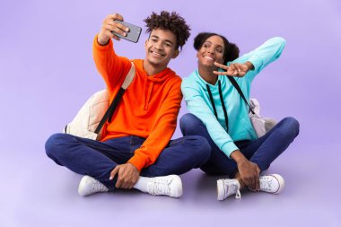 Afro-Amerikan erkek ve kadın bacakları ile katta oturan izole mor arka plan üzerinde çarpı işareti iken selfie fotoğrafı çekmek sırt çantaları giyiyor
