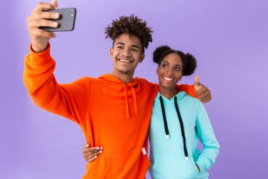 Fotoğraf heyecanlı Afrikalı-Amerikalı çiftin menekşe arka plan üzerinde izole Smartphone cep telefonu ile selfie alarak renkli tişörtü giyen