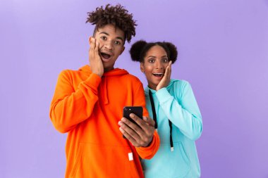 Şok Afro-Amerikan ve cep telefonuyla izole mor arka plan üzerinde renkli tişörtü giyen kız fotoğrafı