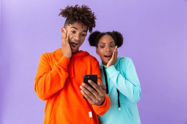 Sürpriz Afro-Amerikan ve cep telefonuyla izole mor arka plan üzerinde renkli tişörtü giyen kız fotoğrafı