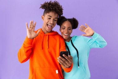 Memnun Afro-Amerikan ve cep telefonuyla izole mor arka plan üzerinde renkli tişörtü giyen kız fotoğrafı