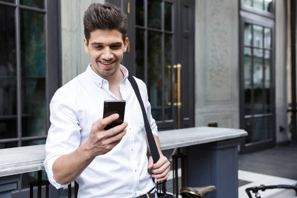 快乐的商人穿着衬衫站在户外的城市街道上 拿着一个袋子 用手机 — 图库照片