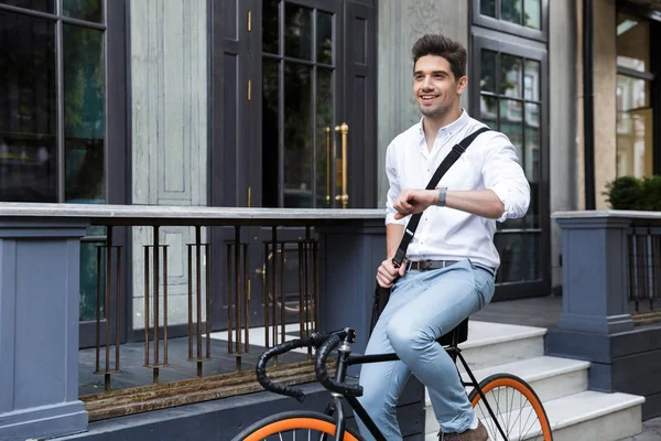 屋外の時間をチェック 袋を持って街で自転車に乗ってのシャツに身を包んだビジネスマンを笑顔 — ストック写真