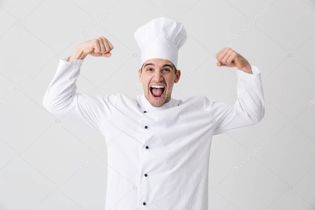 Image of handsome excited young man chef indoors isolated over white wall background showing biceps.