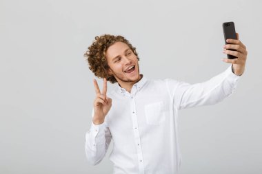 Neşeli bir genç adam cep telefonu ile bir selfie alarak kıvırcık saçlı beyaz arka plan üzerinde izole portresi