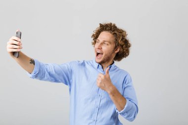 Cep telefonu ile bir selfie alarak mutlu bir genç adam kıvırcık saçlı beyaz arka plan üzerinde izole portresi