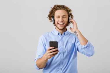 Neşeli bir genç adam cep telefonu tutan kulaklık ile müzik dinlemek kıvırcık saçlı beyaz arka plan üzerinde izole portresi
