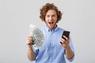 Neşeli bir genç adam cep telefonu tutan para banknot, gösterilen kıvırcık saçlı beyaz arka plan üzerinde izole portresi