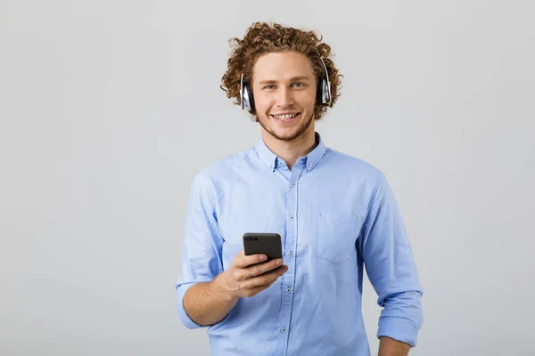 Portrait of a cheerful young man with curly hair isolated over white background, listening to music with headphones, holding mobile phone