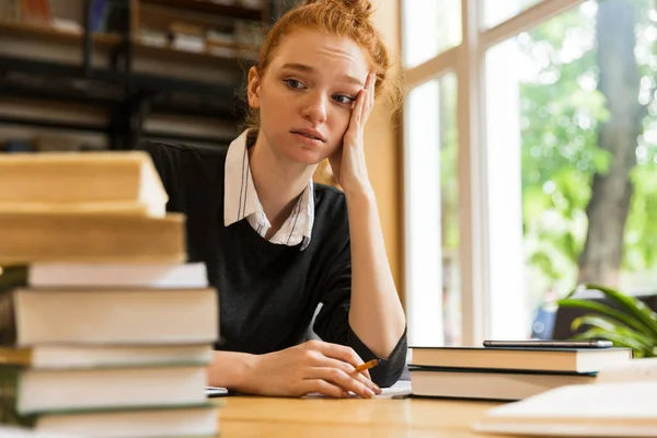 ノートの図書館にある本とテーブルに座って不満混乱して疲れている赤毛の女性生のイメージ — ストック写真