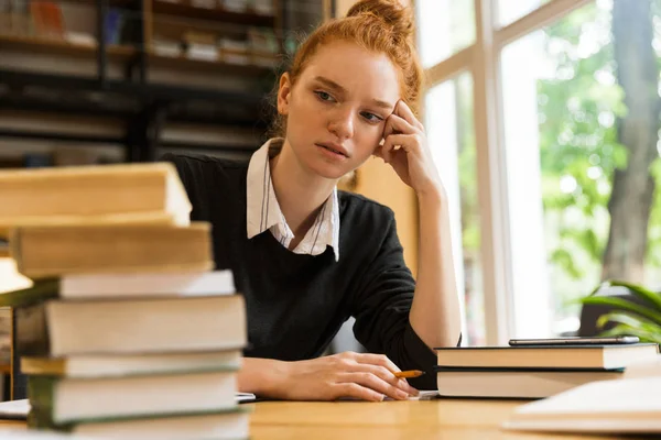不高兴的困惑疲惫的红头发的女学生坐在桌旁的图片与书籍在图书馆写作笔记 — 图库照片