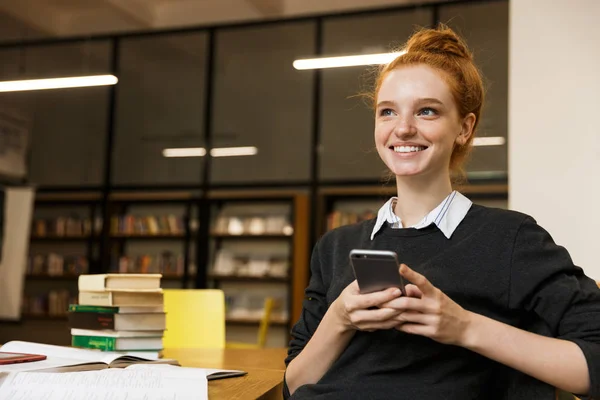 Cep Telefonu Kullaranarak Kütüphane Tabloda Öğrenim Mutlu Kırmızı Saçlı Genç — Stok fotoğraf