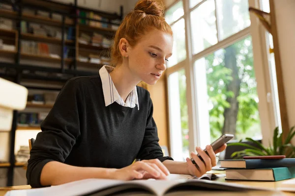 Chica Adolescente Pelirroja Pensativa Que Estudia Mesa Biblioteca Utilizando Teléfono —  Fotos de Stock