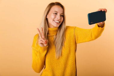 Mutlu bir genç kadının portresi barış gösterilen bir selfie alarak, sarı arka plan üzerinde izole ayakta kazak giymiş