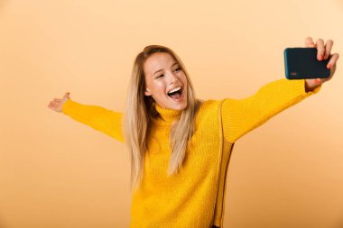Mutlu bir genç kadının portresi bir selfie alarak, sarı arka plan üzerinde izole ayakta kazak giymiş