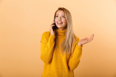 Neşeli bir genç kadının portresi cep telefonu üzerinde konuşurken sarı arka plan üzerinde izole ayakta kazak giymiş