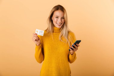 Memnun bir genç kadının portresi kredi kartı, cep telefonu kullanarak gösteren sarı arka plan üzerinde izole ayakta kazak giymiş